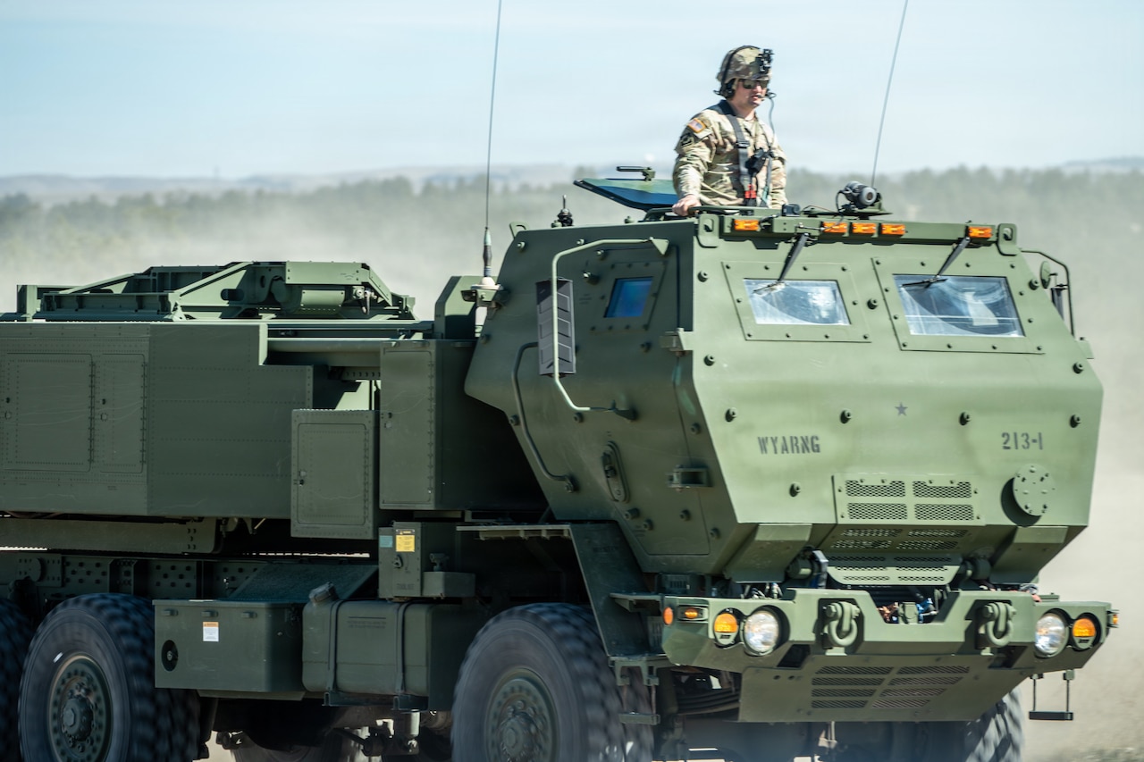 A soldier dressed in a camouflage military uniform stands in the opening of a military vehicle as it drives onto an open field.