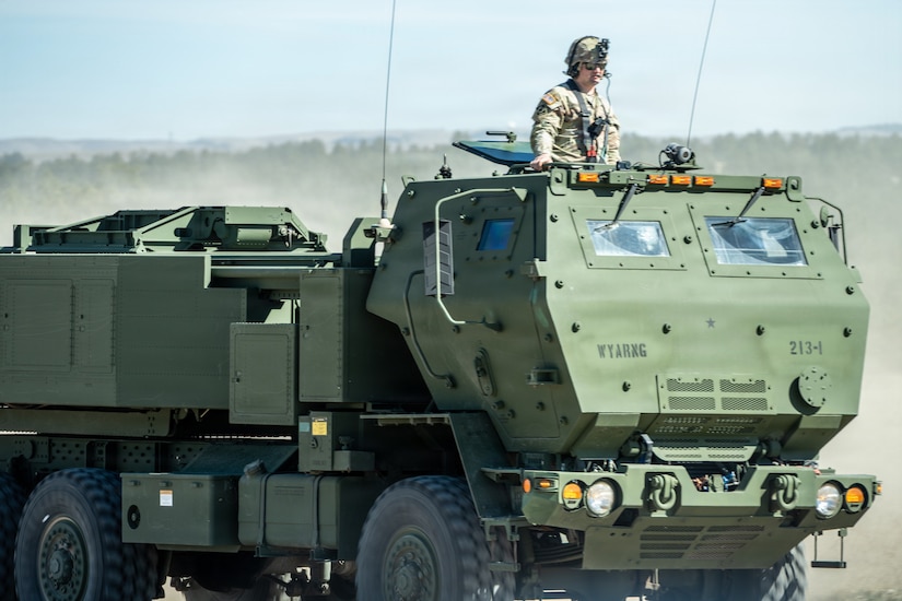 A soldier dressed in a camouflage military uniform stands in the opening of a military vehicle as it drives onto an open field.