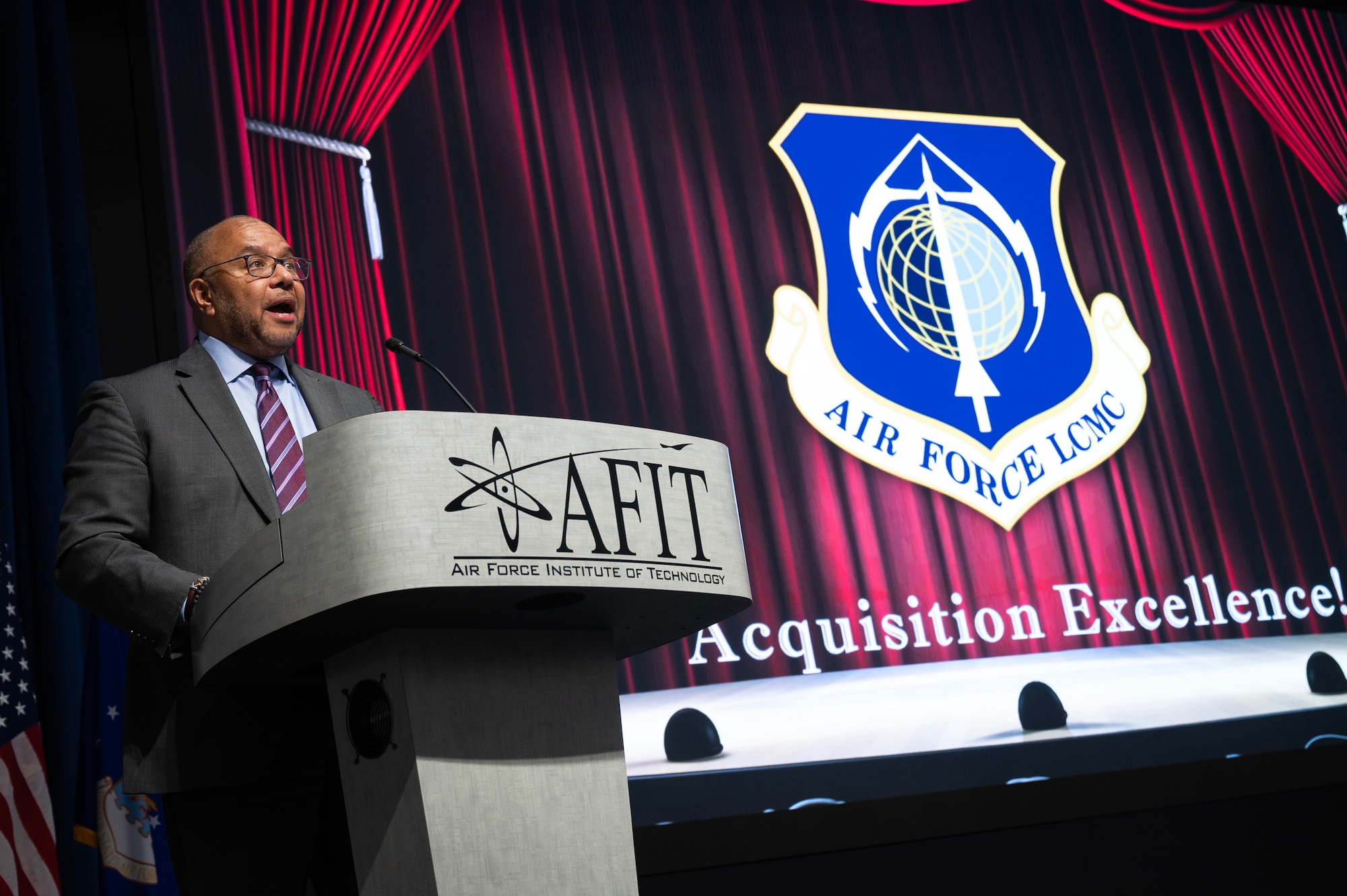 Man stands behind podium. Red curtain is behind him.