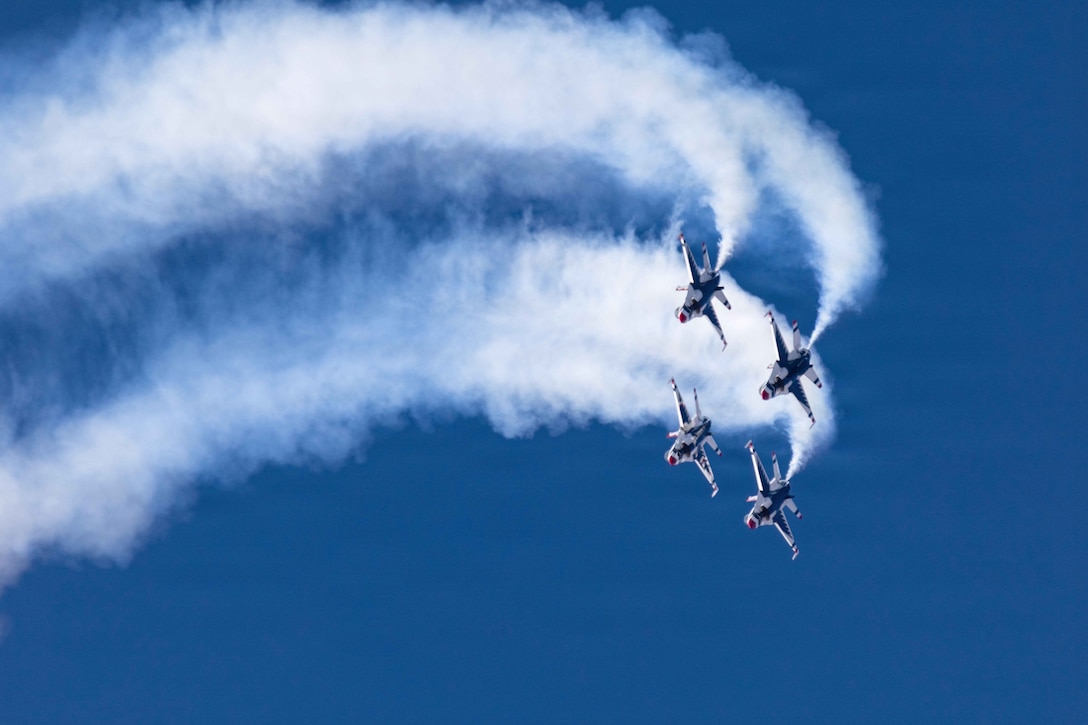 Four aircraft fly in formation in a loop in a blue sky, creating trails of smoke.