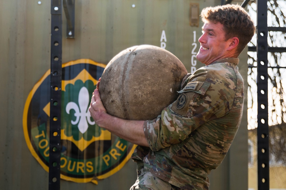 A soldier strains their face while lifting a large stone ball near workout equipment during the day.