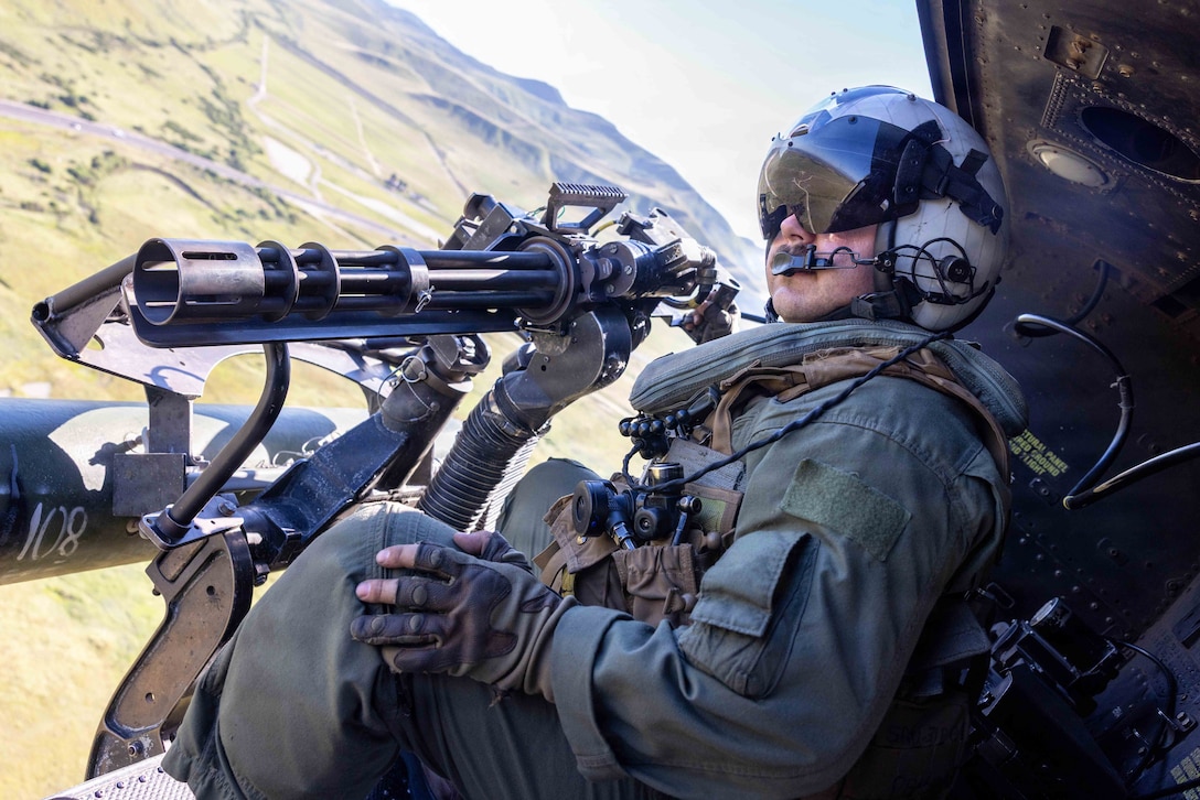 A Marine wearing a helmet sits in the open doorway of a helicopter as it flies in a blue sky over a field with mountains in the background.