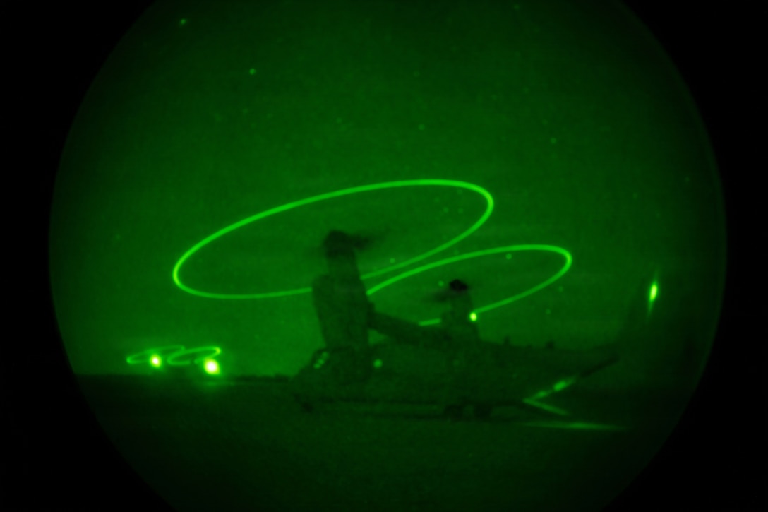 A helicopter sits on a runway in the dark illuminated by green lights as seen through a night vision lens.