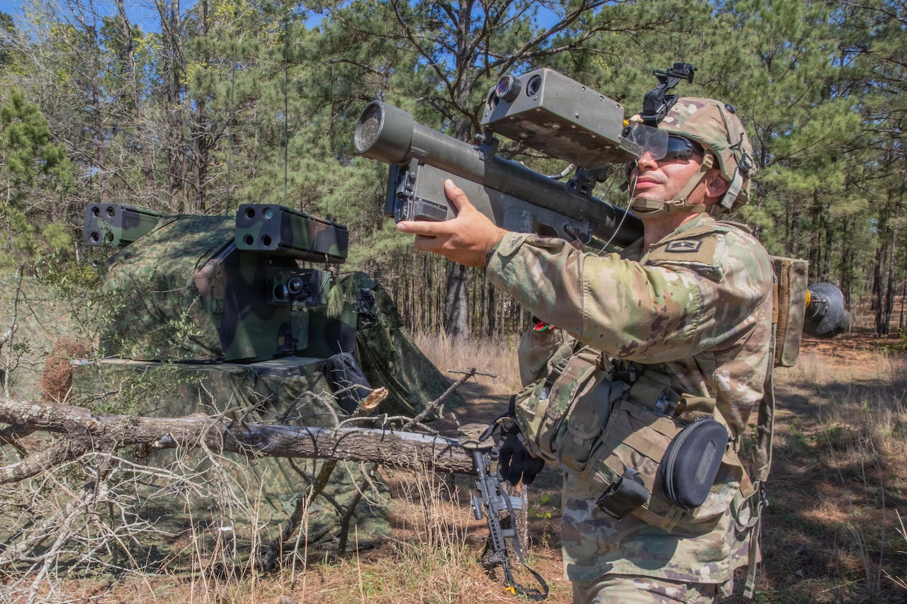 A soldier in tactical gear operates a weapon on their shoulder while standing in front of a wooded area during the day.