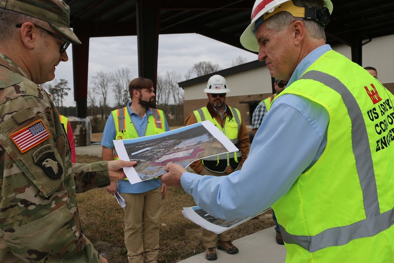 A man in a neon safety vest and helmet holds and points to visuals on an aerieal map, as another man in a U.S. Army uniform looks on.