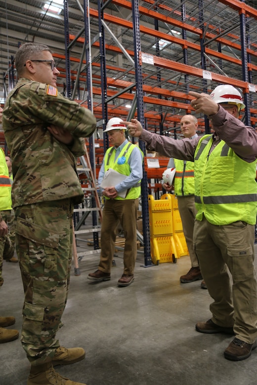 A person in a U.S. Army uniform stands in the left-side of the frame as a person in the right side of the frame in a neon safety vest and helmet points at things. There are heavy-duty shelves in the background.