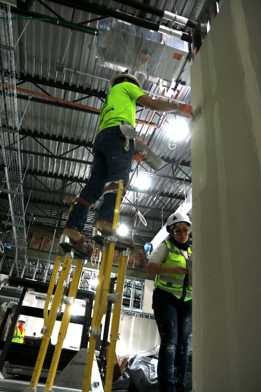 A construction worker stands on stilts and adds compound to unfinished dry walls at a construction site. Another contractor is doing the same from the ground.