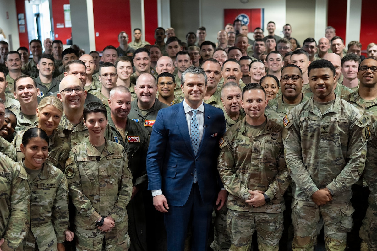 A man in a business suit stands indoors, surrounded by dozens of men and women in military camouflage uniforms, while posing for a photo.