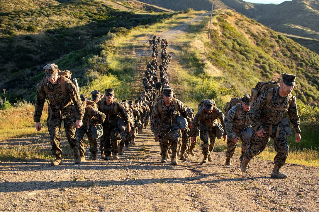 Dozens of Marines walk in a group up a dirt path on a hill.