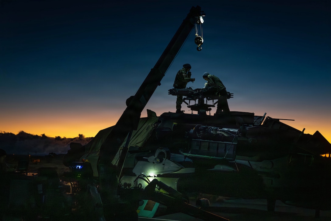 Two soldiers shown in silhouette stand atop a helicopter by its rotor head.