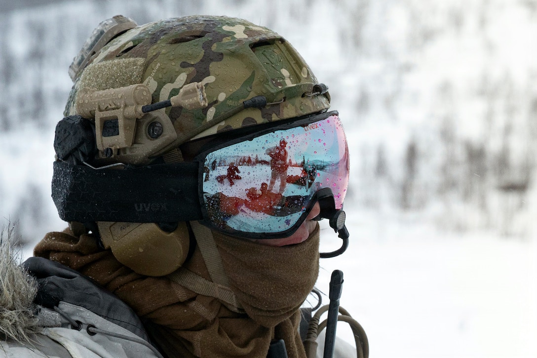 An airman stands in snowy terrain wearing winter gear,  including reflective eyewear that give a view of other troops.