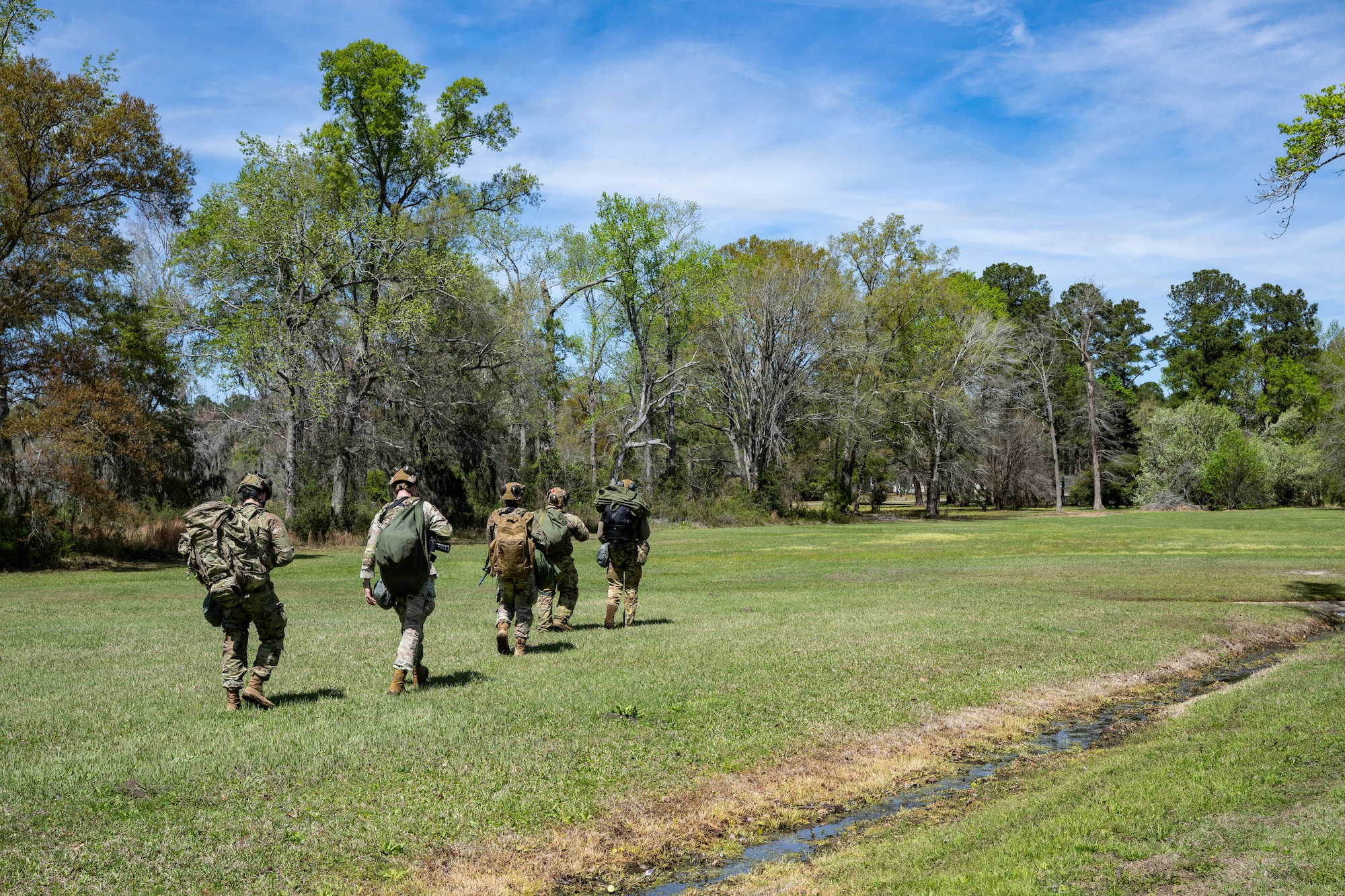 Airmen ruck through a field with bags over their backs