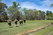 Airmen ruck through a field with bags over their backs