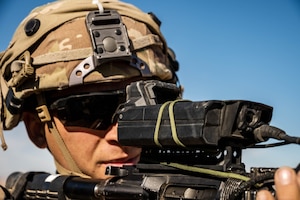 A man wearing a camouflage military helmet and sunglasses looks through a sight mounted on a military weapon outside under a blue sky.
