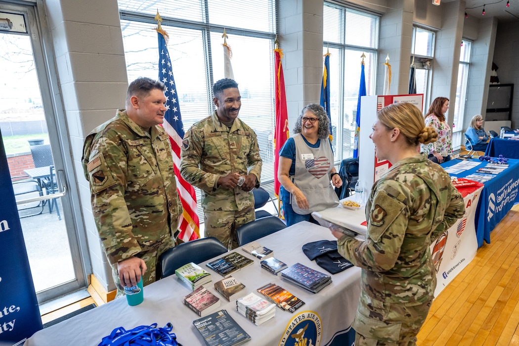 Two men, wearing military camouflage uniforms, and a woman, wearing grey apron, stand behind tables with informational material as a woman, wearing a military uniform stands in front