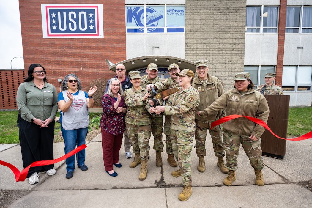 A large group of people, men and women, some military uniforms, other civilian attire, use one large pair of scissors to cut a red ribbon