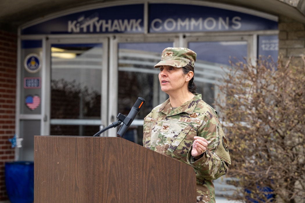 A woman, wearing a military camouflage uniform, stands behind a podium speaking into a microphone