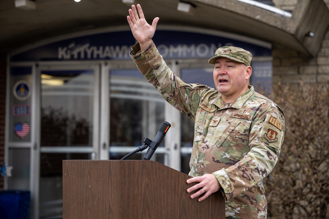 A man, wearing a military camouflage uniform, raises his right hand high as he speaks behind a podium