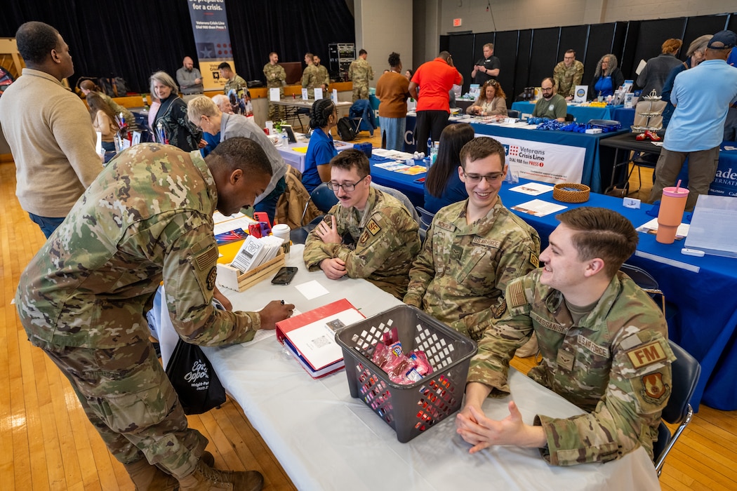 A group of three Airmen sit at a table as another Airmen bends over to sign a piece of paper. Many more people can be seen in the background