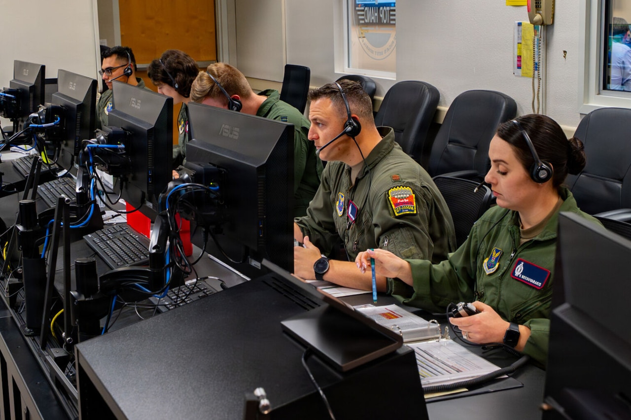 Five people in military flight suits wear headsets and look at computer screens indoors.