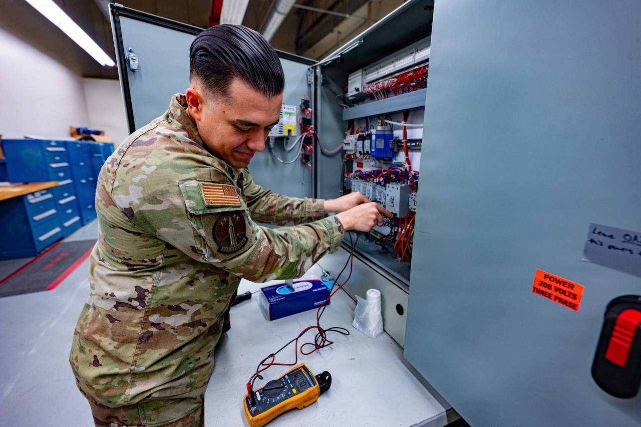 A man in a camouflage military uniform checks a voltage meter while inspecting electrical equipment inside a large room.