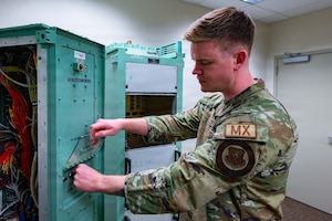 A man in a camouflage military uniform checks electrical equipment inside a room.