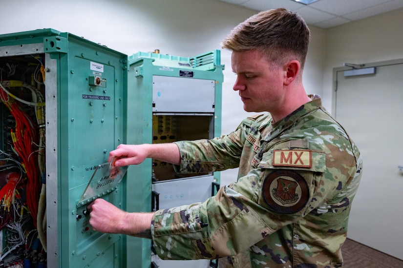 A man in a camouflage military uniform checks electrical equipment inside a room.