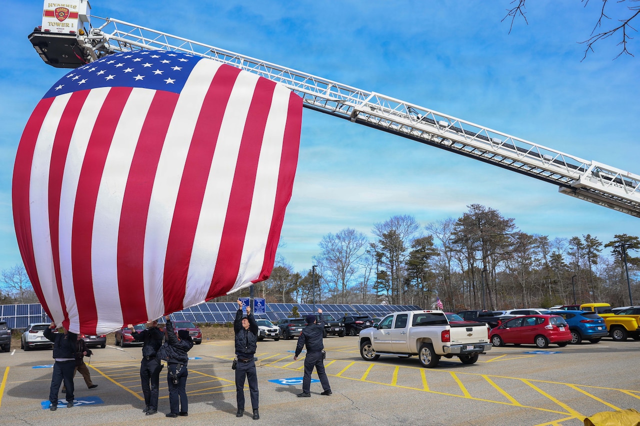 Six men dressed in navy blue jackets and slacks reach up to straighten a large American flag hanging from a crane. Rows of solar panels are nearby, as well as parked cars.