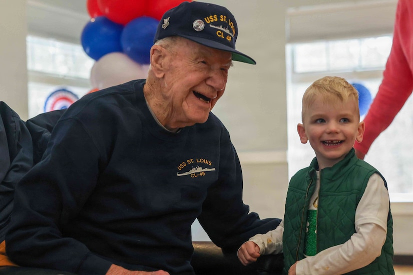 An elderly man wearing a Navy ship memorabilia sweatshirt and cap sits in a black leather chair and smiles. A young blond-haired boy stands next to him and smiles, with other partially visible people nearby. Red, white and blue balloons can be seen in the background.