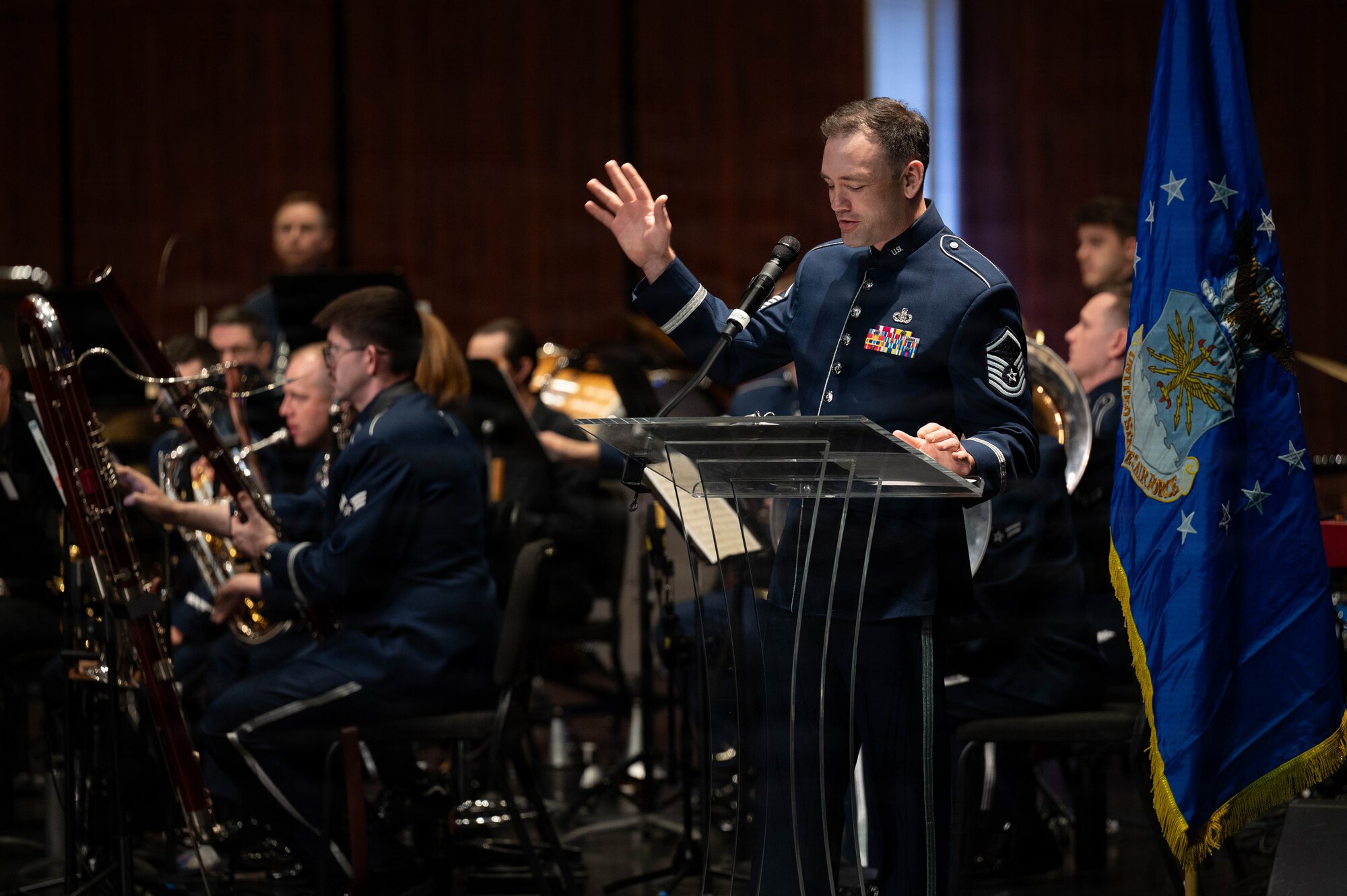 An Airman reads from a script at a podium with band members behind him.