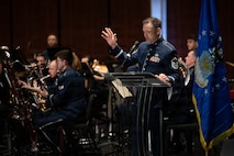 An Airman reads from a script at a podium with band members behind him.