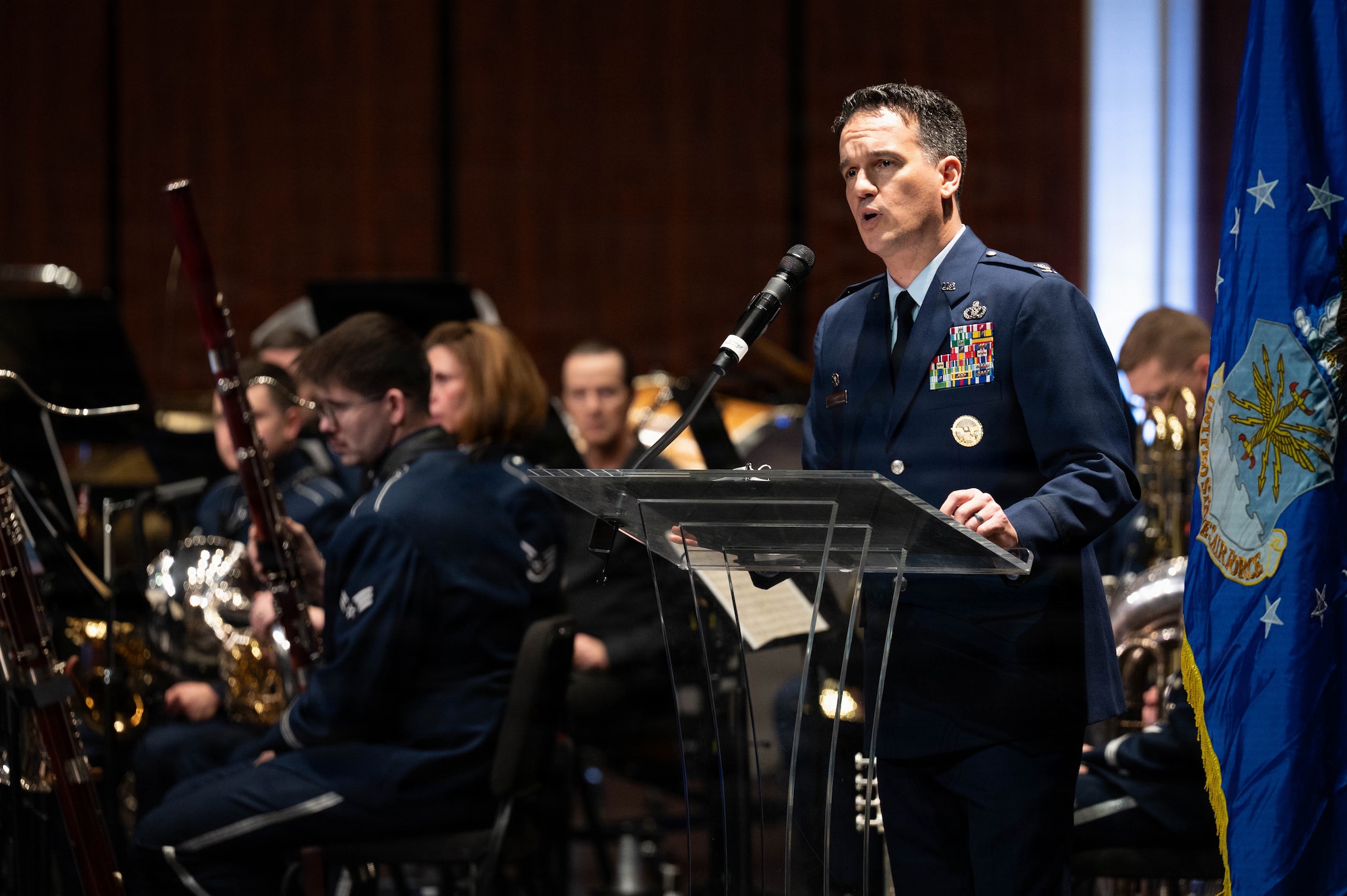 An Airman gives opening remarks for a concert at a podium with band members behind him.