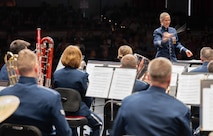 An Airman conducts a band in front of a crowd, the motion of her hand is blurred.