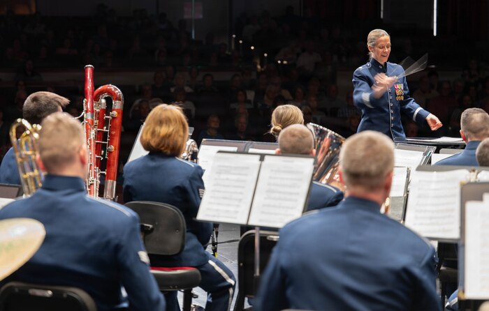 An Airman conducts a band in front of a crowd, the motion of her hand is blurred.