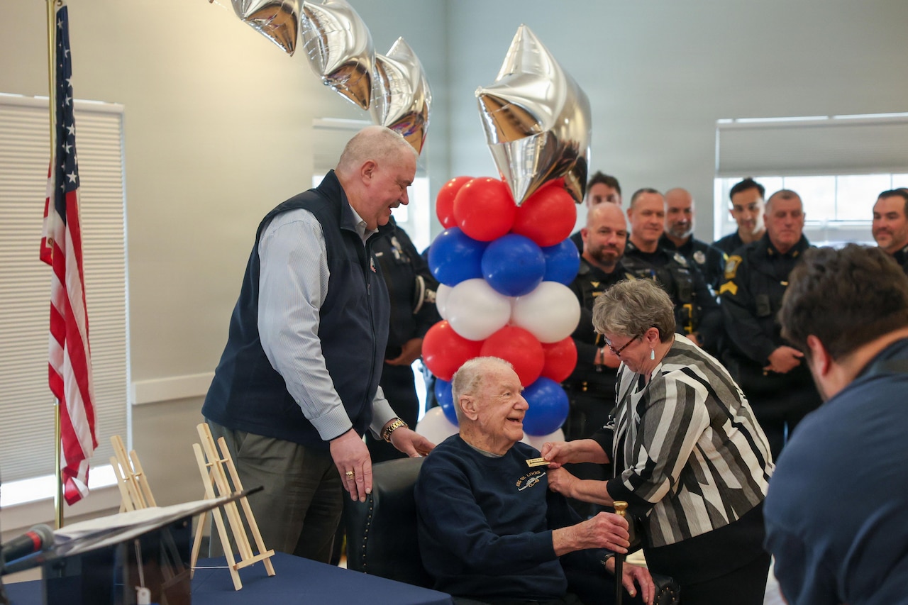 An elderly man sitting in a black leather chair shakes hands with a woman standing in front of him. Other people, some dressed in police and firemen uniforms, stand nearby. A table filled with red, white and blue balloons is behind the man in the chair, where another man is standing.