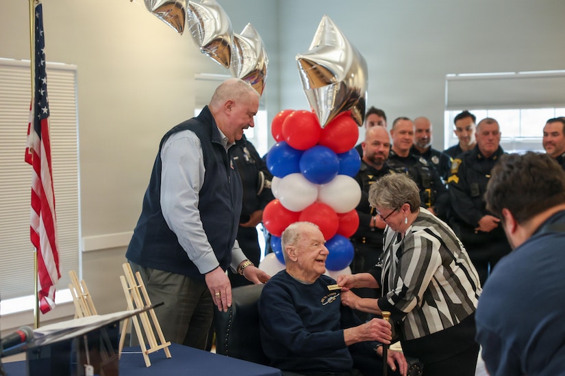 An elderly man sitting in a black leather chair shakes hands with a woman standing in front of him. Other people, some dressed in police and firemen uniforms, stand nearby. A table filled with red, white and blue balloons is behind the man in the chair, where another man is standing.