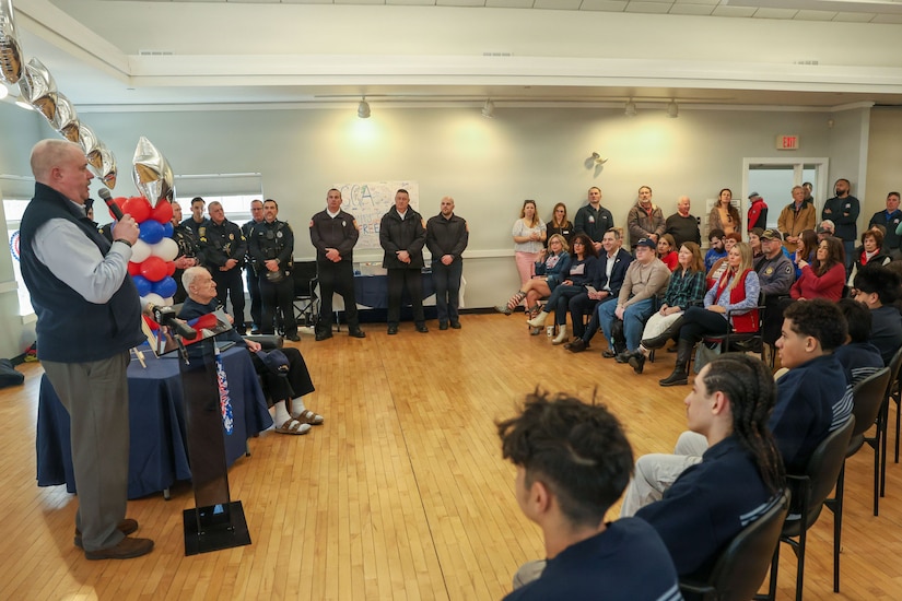 A man holding a microphone stands before a crowd of seated and standing people. Red, white and blue balloons are on a table near the man.