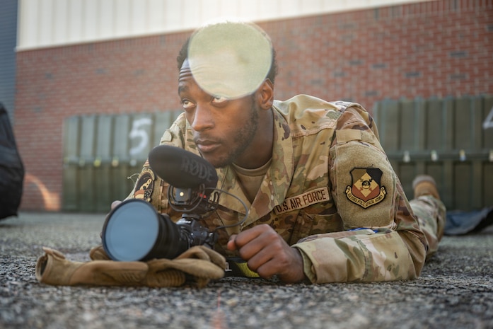 An Airman lays on the ground with a camera resting on a glove in front of him.