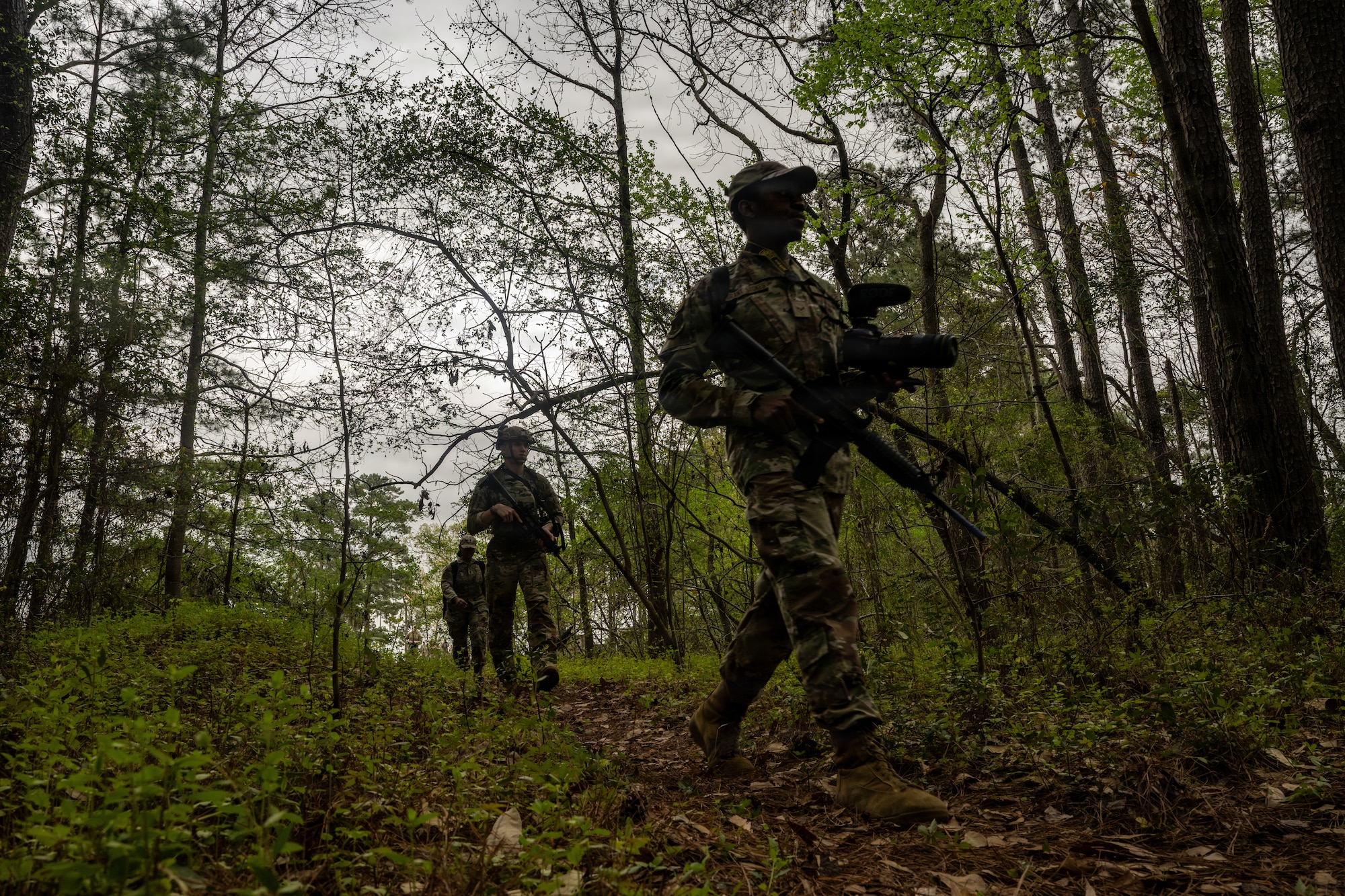 Airmen walk through the woods carrying camera equipment and training weapons.