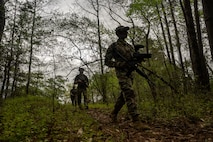 Airmen walk through the woods carrying camera equipment and training weapons.
