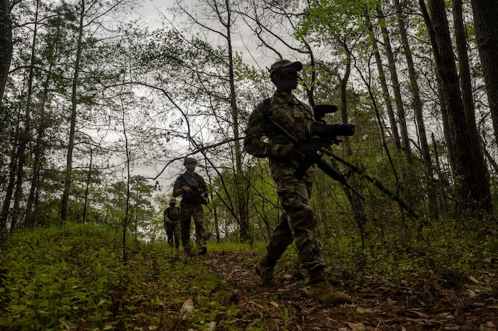 Airmen walk through the woods carrying camera equipment and training weapons.
