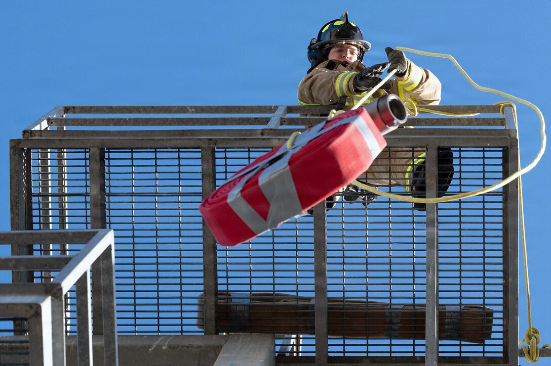 A firefighter, standing on a metal tower and shown from below, pulls up a taped hose roll by a rope.