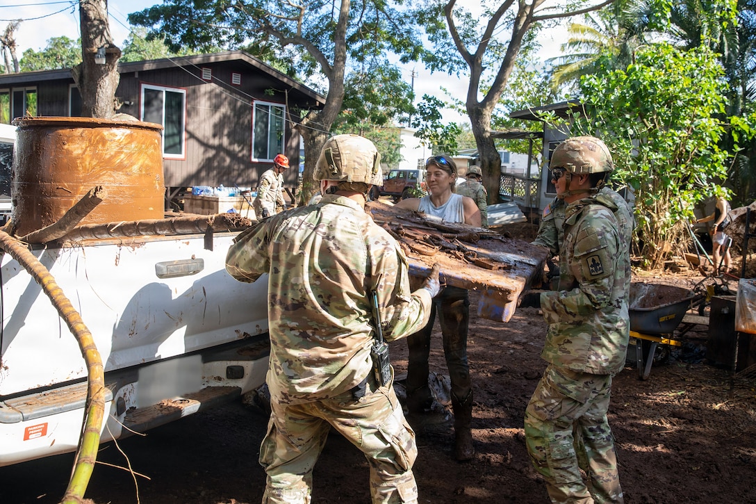 Uniformed service member and a civilian hoist debris into a flatbed truck parked by a house.