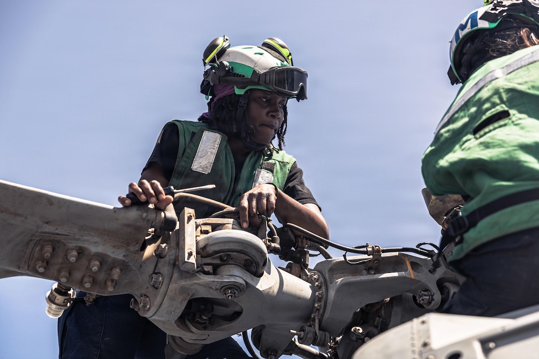 Two sailors in green vests work near a helicopter's blade against a blue sky.