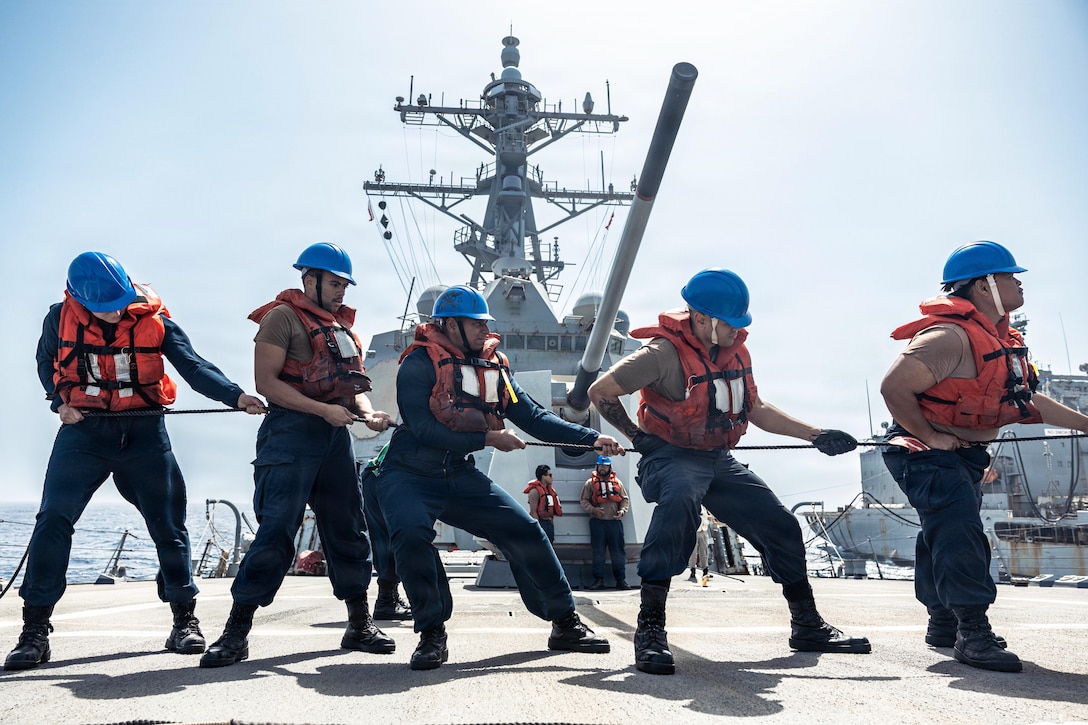 Five sailors in life vests and blue hats hold a line aboard a ship during the day.