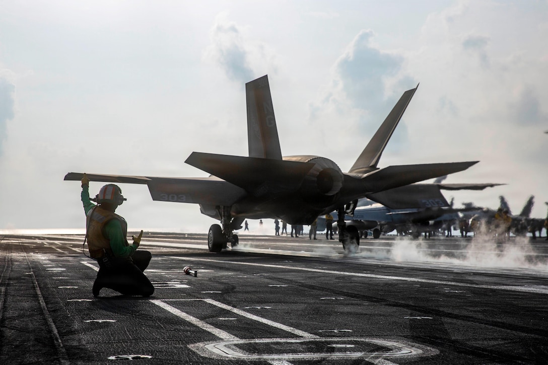 A sailor kneels on a flight deck as an aircraft launches into a bright sky, leaving behind a trail of smoke.
