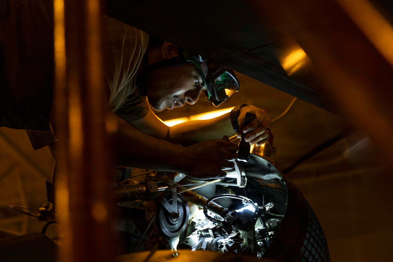 A close-up of a sailor using a flashlight to work on a helicopter in a dimly lit hangar bay.