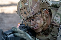 A uniformed soldier in camouflage face paint lies on the ground and looks through a weapon scope with one eye closed.