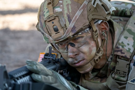A uniformed soldier in camouflage face paint lies on the ground and looks through a weapon scope with one eye closed.