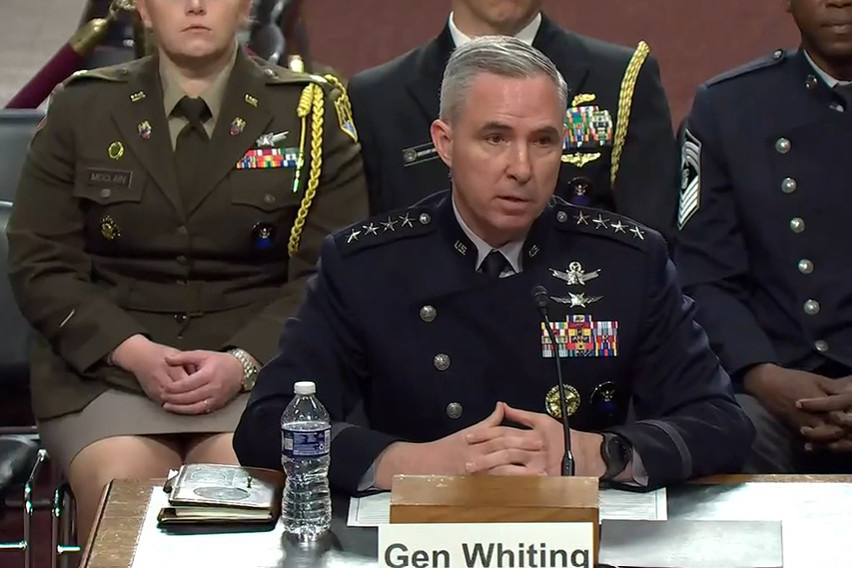 A man wearing a formal military uniform speaks into a microphone while seated at a table. There are people in similar attire seated behind him.